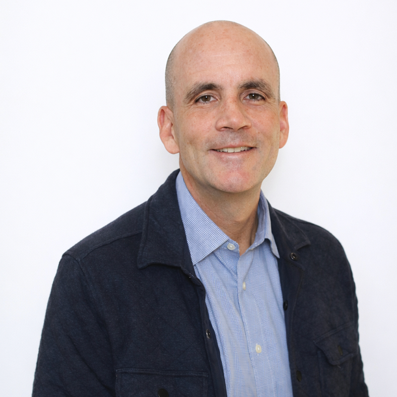 Smiling man in a light blue shirt and dark jacket against a white background (head-and-shoulders).