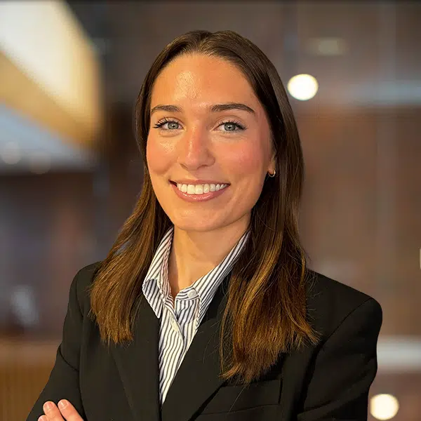 Professional woman in a black blazer and striped shirt smiles at the camera in an office setting.