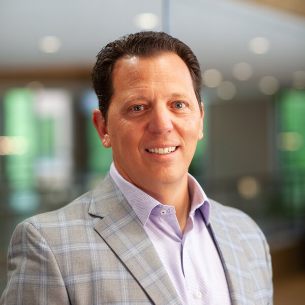 Head-and-shoulders portrait of a smiling man in a gray plaid blazer in a bright office setting.