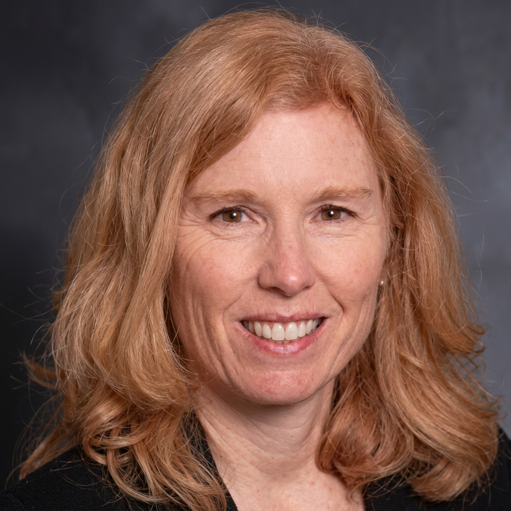 Smiling woman with shoulder-length red hair wearing a dark blazer against a gray backdrop.