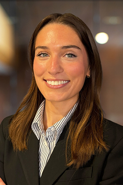 Professional headshot of a smiling woman with shoulder-length brown hair, wearing a black blazer and striped shirt at a neutral background.