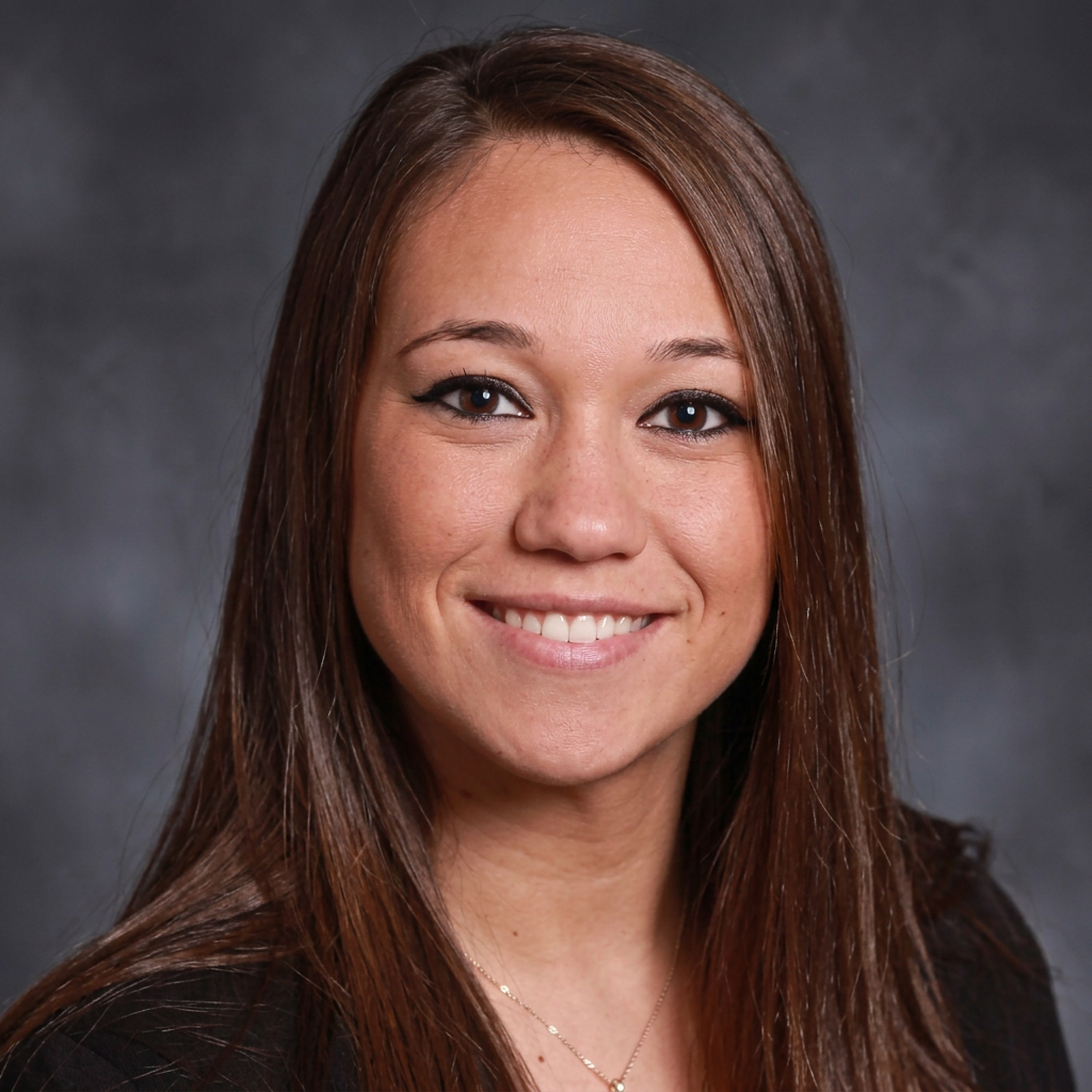 Portrait of a smiling woman with long brown hair wearing a black top, against a gray studio backdrop.
