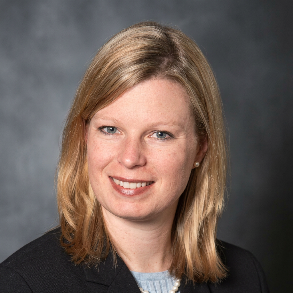 Professional headshot of a smiling blonde woman in a black blazer and light blue top, looking at the camera.